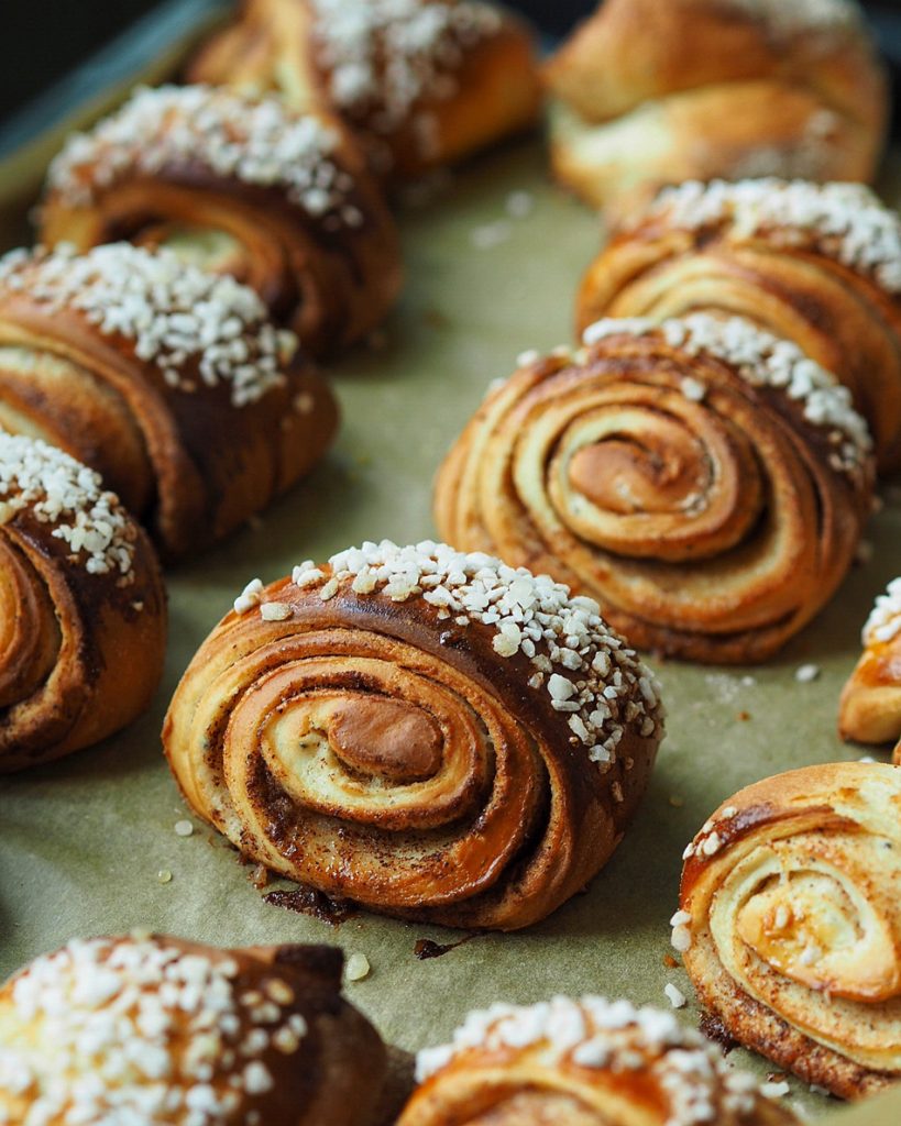 Kuva esittää tuoreita, kanelilla ja raesokerilla koristeltuja korvapuusteja pellillä.
